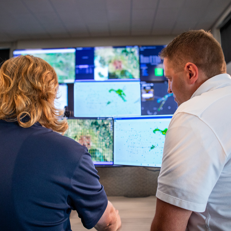 Two people viewing a wall of monitors displaying aerial photographs and weather maps