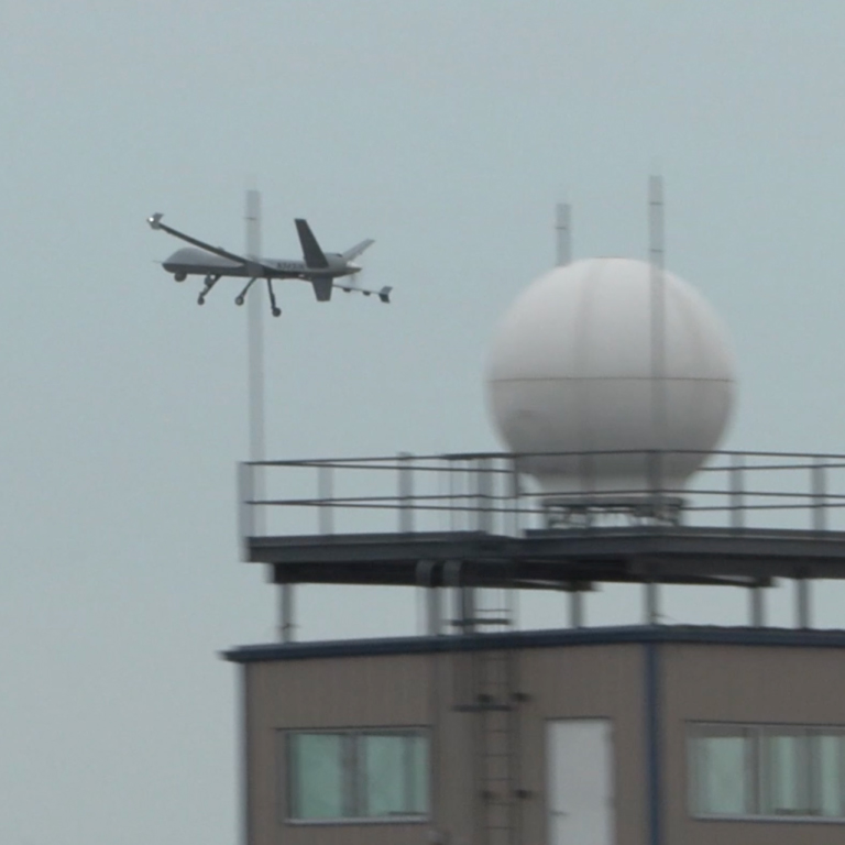 A large unmanned vehicle flying near a building with a radar dome in the foreground