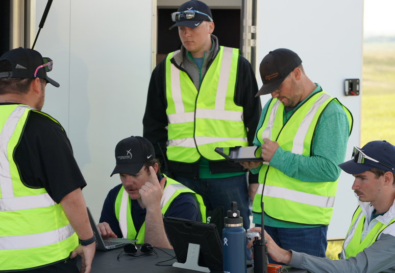 Several men in high-visibility vests are working on laptops and tablets