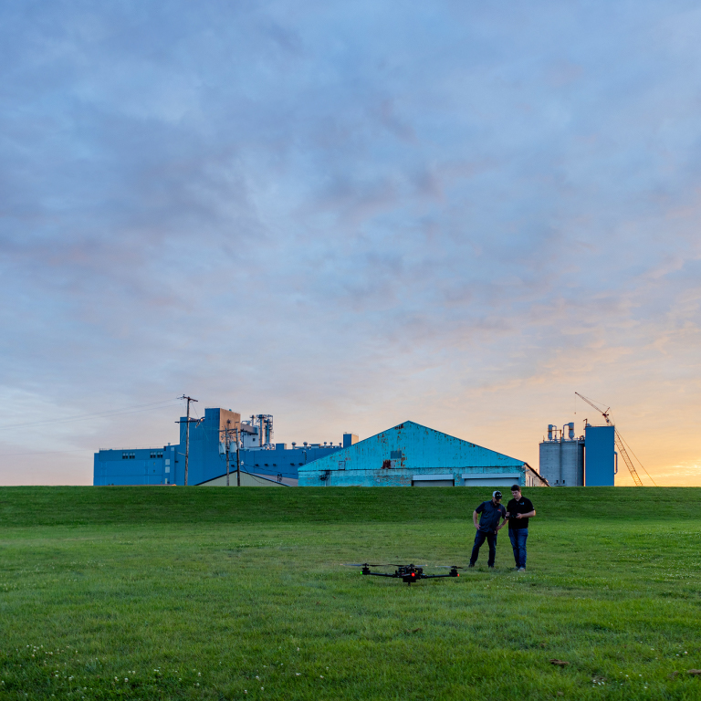 Two men near an unmanned vehicle in a grassy area in front of distant metal buildings